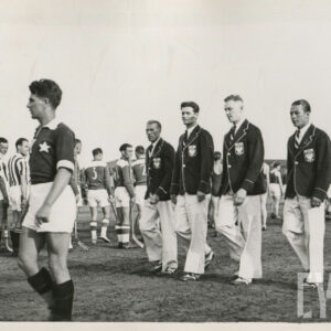 grupa młodych mężczyzn maszerująca po boisku sportowym, część nich nosi eleganckie garnitury okolicznościowe, część stroje sportowe