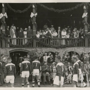 grupa mężczyzn w strojach sportowych, trzymających laski hokejowe, zmierzająca do wyjścia z boiska znajdującego się pod ceglano-drewnianą trybuną, na której stoją oklaskujący ich, elegancko ubrani widzowie