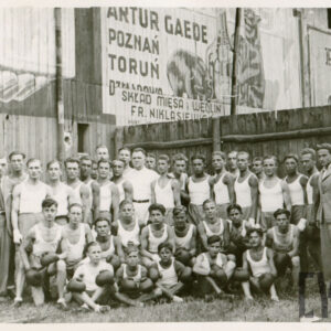 grupa mężczyzn w strojach sportowych, część z nich nosi rękawice bokserskie, część ma zabandażowane dłonie, mężczyźni pozują na tle drewnianego płotu, na którym widać reklamy