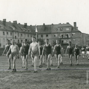 grupa mężczyzn w strojach sportowych, część z nich nie nosi koszulek, maszerująca po boisku sportowym otoczonym drewnianym płotem, w głębi widać ceglane budynki mieszkalne