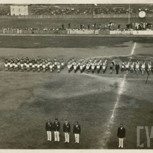 duża grupa osób w strojach sportowych stojąca na boisku piłkarskim, otoczonym trybunami, w głębi widoczne ceglane budynki mieszkalne