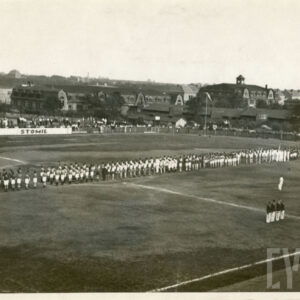 duża grupa osób w strojach sportowych stojąca na boisku piłkarskim, otoczonym trybunami, w głębi widoczne ceglane budynki mieszkalne