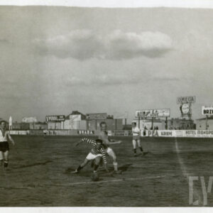 mężczyźni w strojach sportowych, grający w piłkę na boisku otoczonym trybuną z reklamami, w głębi ceglane budynki mieszkalne