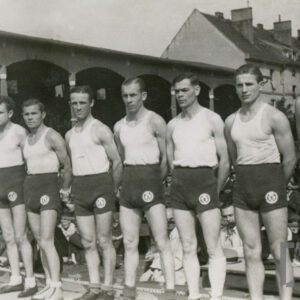 grupa mężczyzn w strojach sportowych stojąca w ringu; w tle widoczne drewniane trybuny i fragmenty domów mieszkalnych