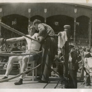 dobrze zbudowany mężczyzna w stroju sportowym i rękawicami bokserskimi na dłoniach, siedzący na taborecie ustawionym w ringu bokserskim, obok niego stoi mężczyzna w średnim wieku; w głębi widać drewnianą trybunę stadionu, na której siedzą liczni widzowie