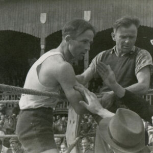 dobrze zbudowany mężczyzna w stroju sportowym i rękawicami bokserskimi na dłoniach stojący w ringu obok mężczyzny w średnim wieku; przyjmujący gratulacje od widzów; w głębi widać drewnianą trybunę stadionu, na której siedzą liczni widzowie