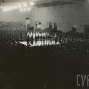 tłum osób otaczających ring ustawiony pośrodku dużej hali; w ringu stoją zawodnicy ubrani w stroje sportowe