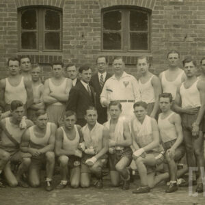 grupa mężczyzn w strojach sportowych, kilku z nich ubranych elegancko, pozujących do fotografii na tle ceglanego budynku