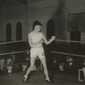 młody mężczyzna w stroju sportowym, stojący w ringu i pozujący do fotografii w pozycji bokserskiej, wokół ringu zebrani są widzowie