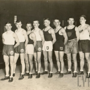 mężczyźni w sportowych strojach, część z nich ma widoczne na twarzy ślady walki, pozujący do fotografii na ringu ustawionym pośrodku dużej hali i otoczonym przez tłum widzów