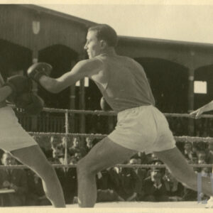 dwóch młodych mężczyzn w strojach sportowych i rękawicach na dłoniach walczących na ringu otoczonym linami, walce przygląda się sędzia w głębi drewniana trybuna, na której siedzą licznie zgromadzeni widzowie