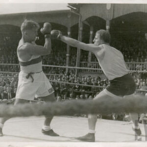 dwóch mężczyzn w strojach sportowych, z rękawicami bokserskimi na dłoniach, walczących w ringu otoczonym linami, w głębi drewniana trybuna stadionu, na której zasiada liczna publiczność