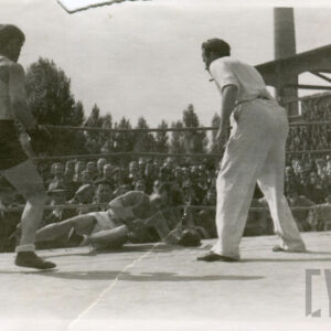 dwóch mężczyzn w strojach sportowych, z rękawicami bokserskimi na dłoniach, jeden z nich leży na macie ringu, drugi stoi nad nim w towarzystwie sędziego, w głębi drewniana trybuna stadionu, na której zasiada liczna publiczność