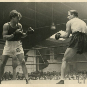 dwóch mężczyzn w strojach sportowych, z rękawicami bokserskimi na dłoniach walczących ringu otoczonym linami, stojącym wewnątrz hali wokół ringu licznie zgromadzona publiczność