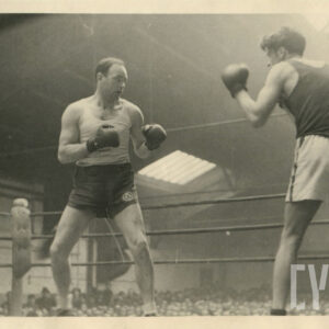 dwóch mężczyzn w strojach sportowych, z rękawicami bokserskimi na dłoniach walczących w ringu otoczonym linami, stojącym wewnątrz hali wokół ringu licznie zgromadzona publiczność