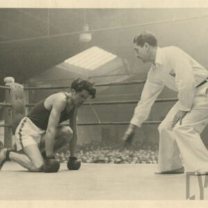 mężczyzna w stroju sportowym, z rękawicami bokserskimi na dłoniach kucający w ringu otoczonym linami, obok niego stoi odliczający czas sędzia, wewnątrz hali wokół ringu licznie zgromadzona publiczność