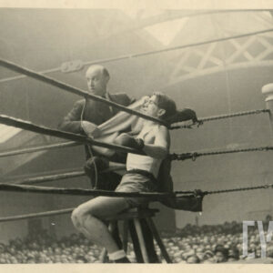 jeden mężczyzna w stroju sportowym, z rękawicami bokserskimi na dłoniach, siedzący na taborecie postawionym w narożniku ringu otoczonego linami, obok stoi mężczyzna w średnim wieku wachlujący ręcznikiem, wokół ringu licznie zgromadzona publiczność