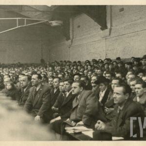 grupa osób, w większości mężczyzn, siedzących w rzędach ławek ustawionych wewnątrz hali wokół ringu