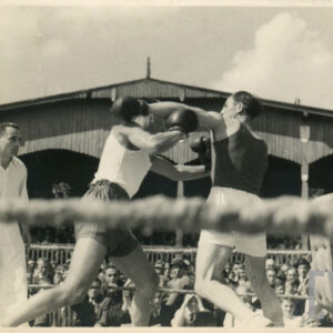 dwóch mężczyzn w strojach sportowych, z rękawicami bokserskimi na dłoniach walczących w ringu otoczonym linami, w głębi drewniana trybuna stadionu, na której siedzi duża grupa widzów
