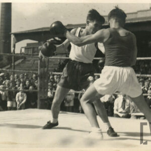 dwóch mężczyzn w strojach sportowych, z rękawicami bokserskimi na dłoniach walczących w ringu otoczonym linami, w głębi drewniana trybuna stadionu, na której siedzi duża grupa widzów