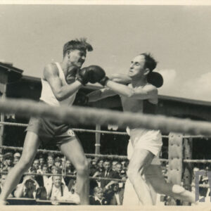 dwóch mężczyzn w strojach sportowych, z rękawicami bokserskimi na dłoniach walczących w ringu otoczonym linami, w głębi drewniana trybuna stadionu, na której siedzi duża grupa widzów