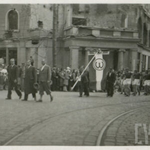 grupa ludzi w strojach sportowych maszerująca za sztandarem z herbem klubu, wzdłuż trasy przemarszu stoi grupa widzów, na drugim planie zniszczony budynek z widocznymi kolumnami