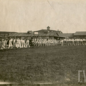 grupa osób ubranych w stroje sportowe, maszerująca bieżnią wokół trawiastego boiska, w głębi drewniane budynki