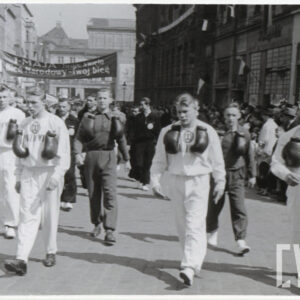 grupa młodych mężczyzn w ubraniach sportowych z przewieszonymi na szyi rękawicami bokserskimi, mężczyźni maszerują ulic, wzdłuż której stoją kamienice