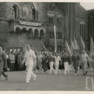 grupa młodych mężczyzn i kobiet w strojach z klubowych i reprezentacyjnych, część z nich niesie w rękach flagi, wzdłuż trasy przemarszu stoi tłum gapiów, w głębi monumentalny budynek z piaskowca
