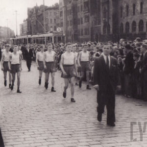 grupa młodych mężczyzn w ubraniach sportowych maszerująca ulicą, wzdłuż której stoją monumentalne budynki oraz tłum gapiów, w głębi widać fragment tramwaju linii nr 3