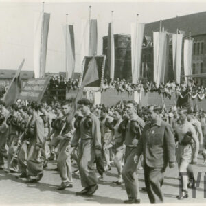 grupa młodych mężczyzn w strojach z klubowych z przewieszonymi przez szyję rękawicami bokserskimi, część z nich niesie w rękach sztandary, wzdłuż trasy przemarszu stoi trybuna honorowa, w głębi monumentalny budynek z piaskowca