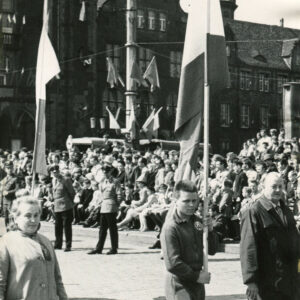 młody mężczyzna w stroju klubowym, trzymający sztandar, maszerujący w towarzystwie starszego mężczyzny i starszej kobiety, w głębi widoczny tłum gapiów lampa uliczna przyozdobiona biało-czerwonymi flagami, w głębi monumentalny budynek z cegły