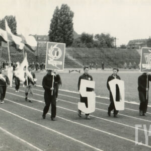 grupa osób w strojach klubowych, maszerująca po bieżni stadionu, trzymająca w dłoniach flagi, sztandary i transparenty
