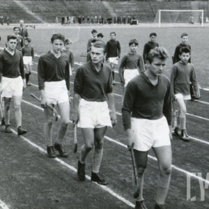 grupa młodych mężczyzn w strojach sportowych, niosąca w dłoniach laski hokejowe, maszerująca po bieżni stadionu