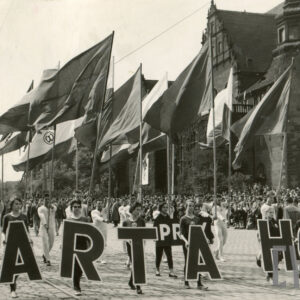 grupa osób w sportowych strojach klubowych, trzymających sztandar, maszerujących brukowaną ulicą, wzdłuż trasy pochodu stoi tłum gapiów, w głębi monumentalny budynek z cegły