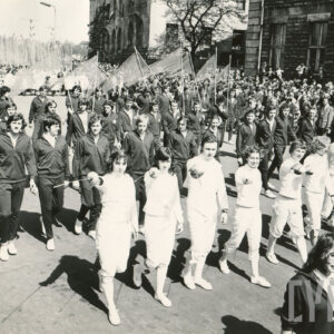 grupa osób w sportowych strojach klubowych, kilkoro trzyma w rękach floret; maszerujących ulicą, wzdłuż trasy pochodu stoi tłum gapiów