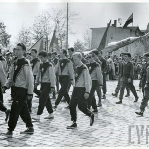 grupa osób w klubowych strojach sportowych maszerująca brukowaną ulicą, część z nich niesie sztandary i flagi klubowe
