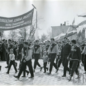 grupa osób w klubowych strojach sportowych maszerująca brukowaną ulicą, dwie osoby niosą transparent z hasłem propagandowym, kilka innych osób niesie sztandary i flagi klubowe