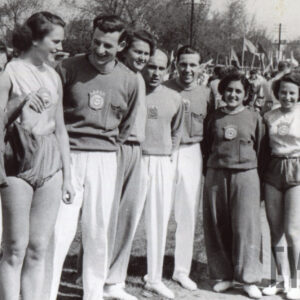 grupa osób - kobiet i mężczyzn, w klubowych dresach, pozująca do fotografii na ulicy, w głębi tłum osób z flagami