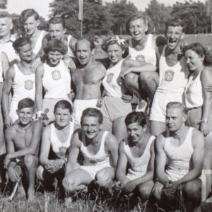 duża grupa osób - kobiet i mężczyzn, w klubowych strojach sportowych, pozująca do fotografii na boisku sportowym, w głębi drzewa i krzaki