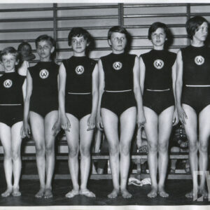 grupa dziewcząt ubranych w stroje gimnastyczne, stojących w rzędzie na hali sportowej; w głębi na ścianie widoczne drabinki sportowe