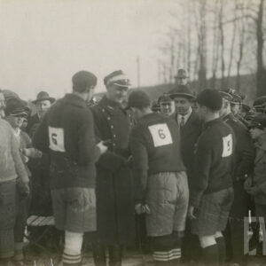tłum mężczyzn, część w strojach do gry w hokeja na lodzie, część w ubraniach codziennych, jeden mężczyzna nosi mundur Wojska Polskiego