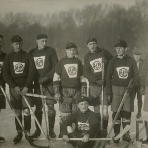 grupa mężczyzn stojących w rzędzie na lodowisku, jeden z nich siedzi na lodzie, mężczyźni ubrani w stroje do gry w hokeja na lodzie, na głowach mają czapki, w dłoniach trzymają koje hokejowe
