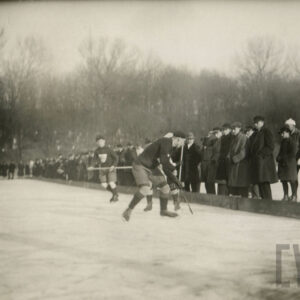 mężczyźni jeżdżący na łyżwach w strojach do gry w hokeja, trzymający w rękach kije hokejowe; obok lodowiska stoi grupa widzów