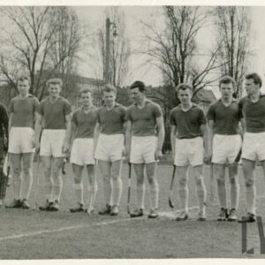 grupa mężczyzn w strojach sportowych stojących w rzędzie na trawiastym boisku, wszyscy trzymają w dłoniach laski hokejowe, a jeden z nich nosi dodatkowe ochraniacze na nogach, w głębi drzewa i fragmenty budynków mieszkalnych