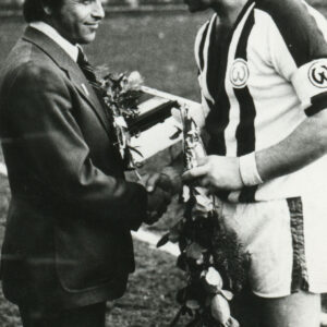 dwóch mężczyzn, jeden ubrany na sportowo składa gratulacje poprzez uściśnięcie dłoni mężczyźnie w garniturze, obaj trzymają w rękach kwiaty