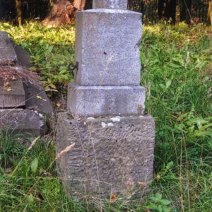 wysoki, kamienny cokół bez krzyża; za nim inny nagrobek, metalowy płot i pnie drzew, teren porośnięty trawą