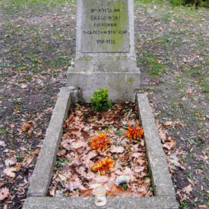 obmurowany grób z pionową stelą na cokole; na tablicy epitafijnej czarne napisy; z tyłu inne nagrobki i pnie drzew