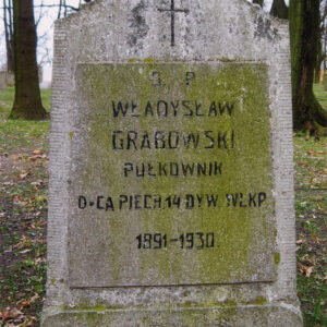 kamienna stela z omszoną tablicą epitafijną, powyżej której znajduje się krzyż łaciński; za nią pnie drzew 