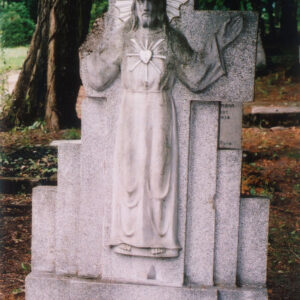 na wąskim cokole znajduje się krzyż łaciński z płaskorzeźbą Chrystusa i schodkowo ustawione kamienne płyty; za nagrobkiem drzewa